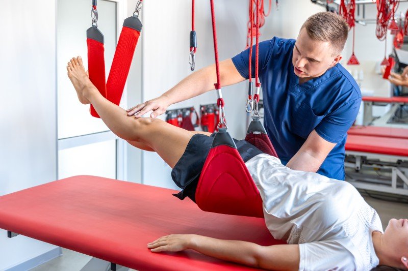 Physical therapist assisting a patient with leg rehabilitation using red suspension straps on a therapy table.