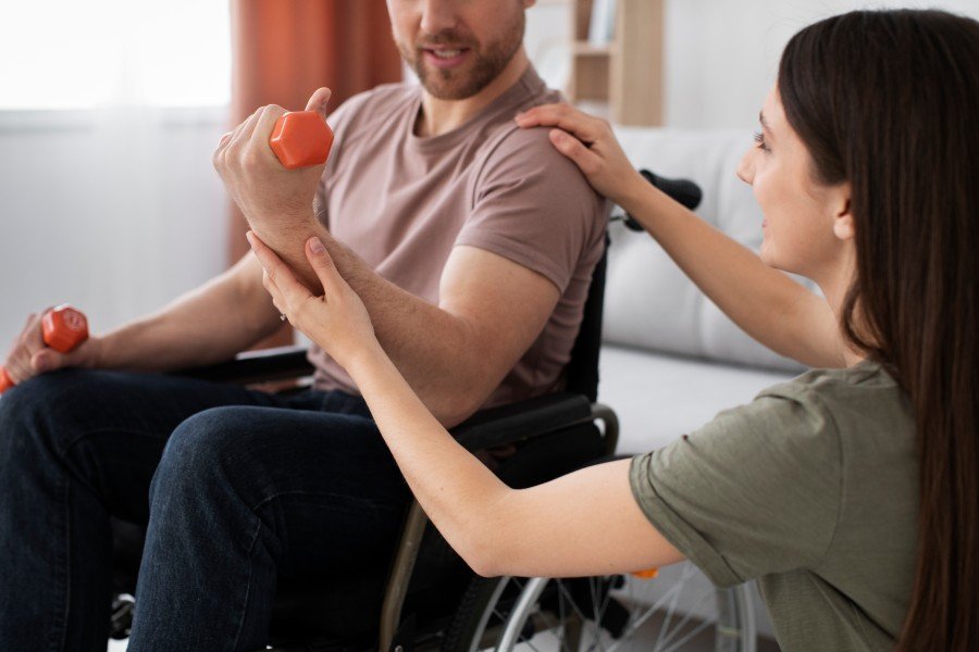 Physical therapist assisting a man in a wheelchair with arm exercises using small dumbbells.